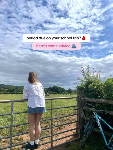 A young woman stands with her back to the camera, leaning on a metal fence. She's facing a green field under a cloudy sky. A bicycle is next to her, and text overlays suggest a video about menstruation.