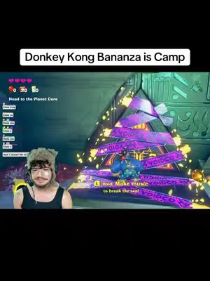 The image shows a man in a fur hat and glasses looking at the camera. The screen behind him displays gameplay of a video game. There's text overlay that says  'Donkey Kong Bananza is Camp.' Below him, there is an overlay of the game interface with information about the game