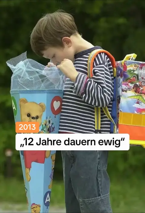 A young boy in a striped shirt and jeans is looking into a large, decorated cone-shaped container, likely a school bag for the first day of school, with a blurred green background. The cone is blue with a teddy bear motif, and the added text says something along the lines of “12 years take forever”.