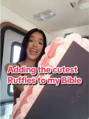 A young woman with dark hair and a gentle smile is holding a large Bible with decorative ruffles on it; she is standing in front of a window, with the text "Adding the cutest Ruffles to my Bible" displayed in a bubble near the bottom of the screen. The background is slightly blurred, and the overall composition creates a sense of homemade charm and creative expression.