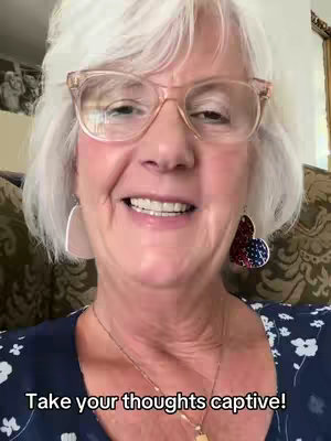 A close-up shot of a light-skinned elderly woman wearing glasses. Her smile is bright, and she is wearing large heart-shaped earrings with a red, white, and blue pattern. The background is blurred, suggesting a casual indoor setting with couch details visible.