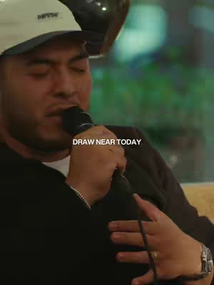 The cover image shows a man in a black jacket and a white cap holding a microphone. The background is blurred, with out-of-focus elements in green and yellow providing subtle color gradients. The text overlay "DRAW NEAR TODAY" is positioned at the bottom center.