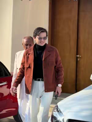 A well-dressed man in sunglasses smiling, standing next to a car. He is wearing a brown jacket, black turtleneck, and white pants. Another man in a white suit is partially visible behind him. The background includes a building and a wooden door.