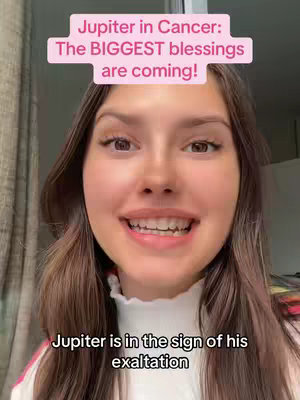 A selfie-style shot features a woman with long brown hair. She is centered in the frame, smiling and looking directly at the viewer. Above her head, in a bright pink rectangle, is text: "Jupiter in Cancer: The BIGGEST blessings are coming!" At the bottom, the text reads "Jupiter is in the sign of his exaltation". The background is blurred, but appears to be an indoor setting with a window and curtains.