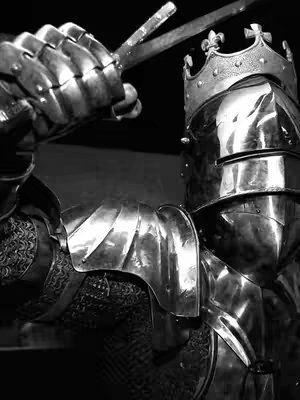 A black and white image of a knight in full armor, viewed from a slightly low angle. The knight's helmet, adorned with a crown, reflects the light. One gauntleted hand holds a sword angled upward. The composition is dramatic, focusing on the details and texture of the armor.