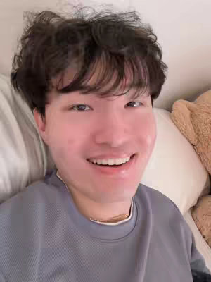 A selfie of a young man smiling broadly while lying down in bed. The lighting is soft, with a light-colored pillow and a fluffy brown teddy bear visible at the edge of the frame. He wears a light blue long-sleeved shirt with a gold chain, with dark hair and a flushed face.