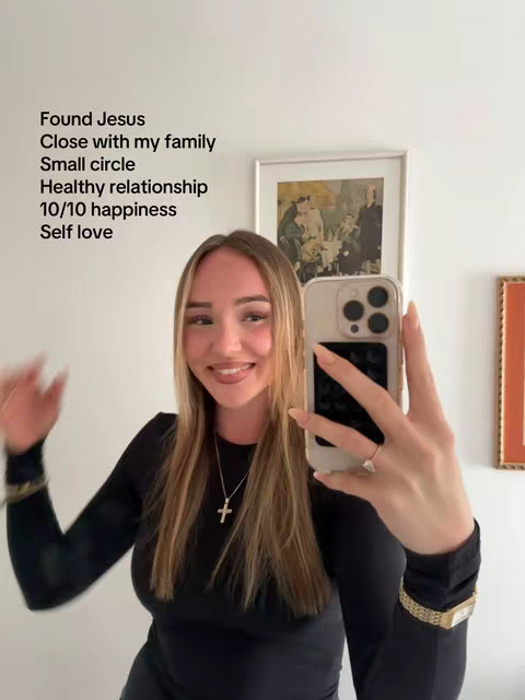 A woman is taking a selfie in a well-lit room. She is smiling at the camera and holding her phone with her right hand. Text overlays are visible, listing items relating to happiness and self-love. In the background, there are framed artworks and beige walls.