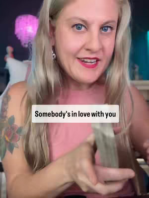 A woman with long blonde hair is centered in the frame, looking towards the viewer and smiling. She's wearing a pink top with a rose tattoo visible on her upper arm. A text overlay reads 'Somebody's in love with you'.