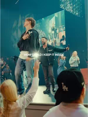 The cover image captures a live worship service. In the foreground, a person in a black cap faces away from the camera, with raised hands. In the middle ground, two male figures—one dressed in jeans and a hoodie appears to be singing, while the other is in a black jacket and is holding a microphone and singing. Behind, a woman sings through the microphone. The setting appears vibrant with bright stage lighting and projected images on a large screen, along with a crowd of people visible in the background.