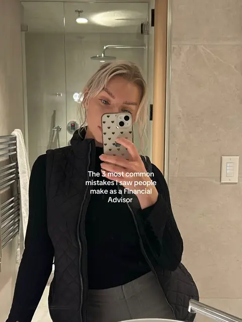 A woman holds a phone, taking a selfie in a bathroom mirror. The background is a modern bathroom with a shower visible. Text overlayed on the image. The woman is wearing a black vest and shirt. The phone has a heart patterned case.
