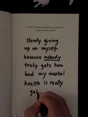 The image shows an open notebook page filled with handwritten text. Above the text is a small heading. In the foreground, a hand holding a pen is partially visible, suggesting the text is being written or emphasized.