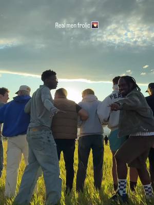 A group of men frolicking in a field at sunset, the sky is filled with clouds, and the sun is setting behind them. The text 'Real men frolic' is overlaid on the top of the image.