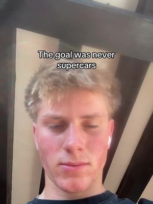 A selfie-style image featuring a young man with fair skin and short, light-colored hair. He is wearing a dark-colored shirt and partially out-of-frame white earbuds are visible. The man's expression is neutral. Text overlayed on the image reads "The goal was never supercars."