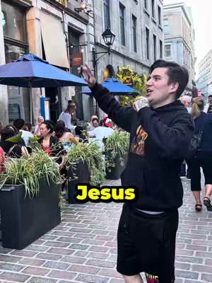 A man wearing a black hoodie and shorts stands on a brick pathway in a city setting, holding his arm out. The man has tattoos on his arm with the word Jesus in a yellow, outlined font at the base of the image.