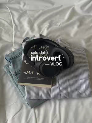 The cover image displays a casual arrangement on a white bedsheet. A pair of blue jeans lies at the bottom left, followed by a folded, striped button-down shirt. A closed book with dark cover and white text is positioned in the center, with a pair of black headphones resting on top. The hook "solo date introvert -VLOG" is superimposed over the headphones element.