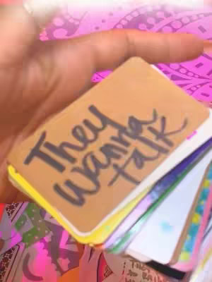 A hand holds a deck of tarot cards, the top card facing the viewer. The card is beige with the text 'They wanna talk' scrawled on it in black ink. Other cards of various colors and designs are visible behind the top card.