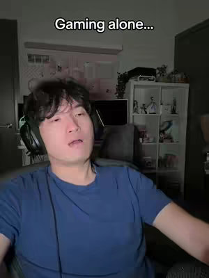 A man is seated in a room, looking at the camera with a neutral expression. He is wearing a blue t-shirt and a headset, suggesting he is gaming or engaging in online activities. The room is somewhat dimly lit, with a shelf and other items visible in the background, indicating a personal living space. Text overlayed on the image reads "Gaming alone..."