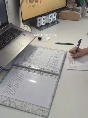 An overhead shot of a desk with a laptop, a digital clock, and study materials. A person's hand is visible, writing on a paper. The setting is well-lit and organized, suggesting a study environment.