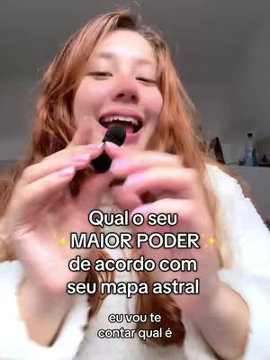 A woman with long reddish-brown hair, holding a small black microphone in front of her mouth while laughing, her face taking up the majority of the vertical frame. The background is blurred but appears to be a room with some light items on a shelf. Overlay text in white lettering and glittery star decorations is centrally placed, with the first line "Qual o seu" at the top, followed by "MAIOR PODER" in larger text, then "de acordo com", and finally "seu mapa astral". Below is "eu vou te", followed by "contar qual é".