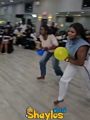 Two women are running while holding balloons in a brightly lit room. Behind them, other people are seated at tables.  The camera is focused on the women, highlighting their actions and expressions.