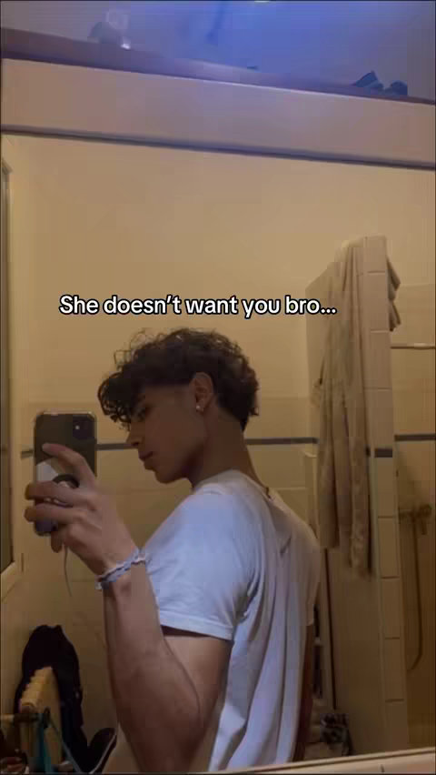 A young man in a white t-shirt is taking a selfie in a bathroom mirror. Text overlayed reads 'She doesn't want you bro...'. The background features tiles and a shower curtain.