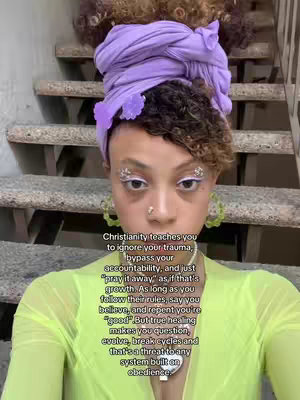 A close-up shot of a person against a backdrop of concrete stairs. The person has their hair styled in a purple turban with a flower pinned on top, and they appear to be looking directly at the camera. They are wearing a light green shirt, with shiny embellishments on their eyes. The background consists of a series of concrete stairs.
