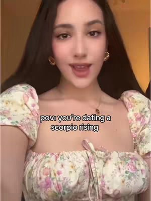 A young woman with long dark hair and wearing a floral top is looking at the camera and speaking. Text overlay states "pov: you're dating a scorpio rising". The background is blurred, with warm lighting.