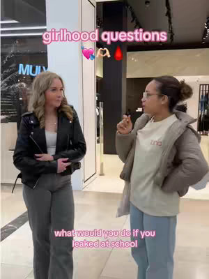 Two young women stand in front of a store entrance in a mall setting, facing each other and talking. Text overlays indicate the subject is about 'girlhood questions'. The woman on the left is wearing a black leather jacket and grey pants. The woman on the right has a puffer jacket over a t-shirt and is wearing glasses. The overall composition captures a casual, conversational style.
