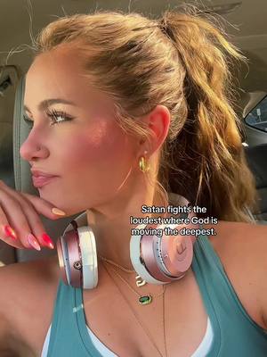 A young woman with light skin and blonde hair, styled in a high ponytail, is shown from the chest up. She is angled slightly towards the left, looking leftward, with a neutral expression. She has visible makeup including blush and lip color. She wears gold hoop earrings and a light makeup palette. Headphones in white and rose gold are hung around her neck. The background suggests she is in a car, with parts of the interior in view. Text overlaying the image states a sentiment about spiritual warfare.