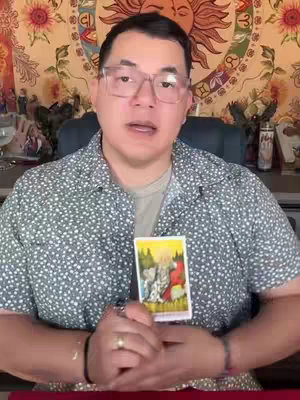 A man with glasses is centered in the frame, holding a tarot card. He's seated in front of a tapestry with a sun design. The background is blurry but suggests a room setting, with items on display.