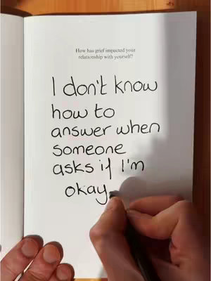 The image shows an open book with handwritten text and a hand holding a pen as if writing. The top of the page reads 'How has grief impacted your relationship with yourself?' below which the following is written: 'I don't know how to answer when someone asks if I'm okay.' The image has a warm, neutral color palette.