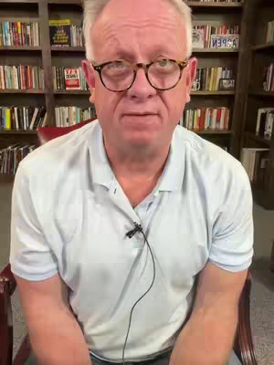 A middle-aged man with glasses, wearing a light blue collared shirt, is seated and looking directly at the camera. Behind him are bookshelves filled with books.