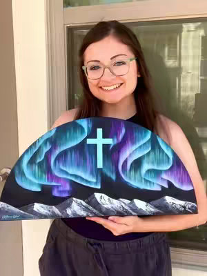 A woman with glasses smiles broadly while holding a painting depicting a stylized aurora borealis scene with a cross in the center. The painting is an arched shape, featuring blue, purple, and white colors representing the northern lights, mountains, and a bright blue cross.