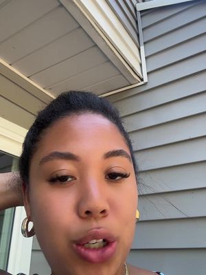 A young woman with dark hair and hoop earrings appears in a selfie style shot. She is angled slightly and is speaking, with a neutral background. The lighting is bright and natural, highlighting her facial features.