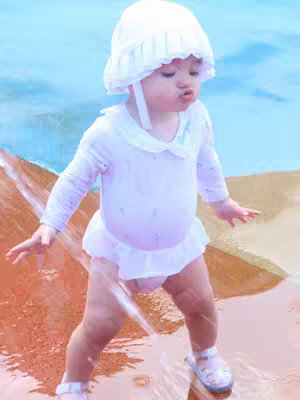 A candid shot captures a toddler girl enjoying water play. She stands in shallow water wearing a white sun hat and a white onesie with a ruffled skirt. Water streams down, creating a playful environment, and she has a surprised expression.