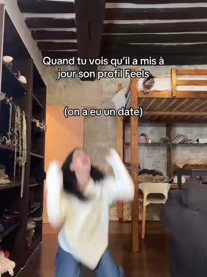 A woman in a white sweater and jeans is joyfully jumping toward the camera. The background includes a room with wooden beams on the ceiling, a dark wooden closet on the left, and a bunk bed and chair on the right, creating a casual, interior setting.