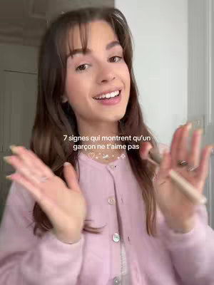 A woman with brown hair and bangs smiles while holding a makeup product in her hand. The background is a soft, neutral tone. The image is well-lit, and the woman appears to be in a room with a neutral color palette.