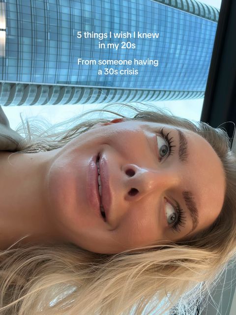 A selfie-style shot featuring a woman looking up at the camera with a slight smile. The background is a blurred view of a modern building with many windows, reflecting the sky. Overlayed text appears at the top, stating '5 things I wish I knew in my 20s' and 'From someone having a 30s crisis' underneath it. The woman has blonde hair and is positioned close to the camera, with her face taking up a large portion of the frame.