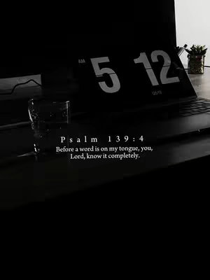 A dark, moody image featuring a laptop and a glass of water on a desk. On the laptop screen, a digital clock displays the time 5:12 AM/PM. Beneath the screen, a Bible verse from Psalm 139:4 is present in white font.