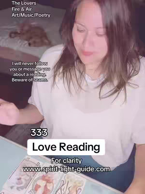 A woman is seated at a table, performing a tarot card reading. The scene is moderately lit, with soft lighting and a blurred background. The woman, with long dark hair and a white top, is looking down at the cards with a focused expression. Text overlays at the top and bottom partially obscure the image.