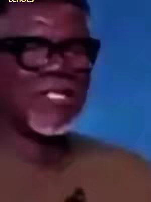 A close-up of a Black man wearing black-rimmed glasses. He appears to be speaking, with his mouth slightly open. The background is a blurry blue.