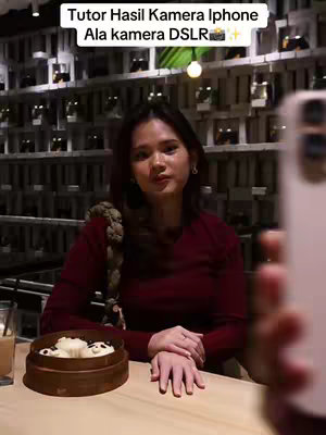 A woman sits at a table, looking at the camera. She has dark hair and is wearing a maroon top. In front of her is a bamboo steamer basket containing food. The background is a restaurant setting with shelves.