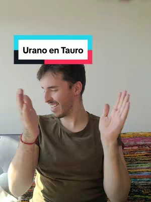 A man with dark hair is the central figure. He's wearing a green t-shirt and has his arms open. Above him, a banner reads 'Urano en Tauro' in white text against a black background, set upon a turquoise and pink stripe background. The background is a plain white wall and the upper portion of an object. The man appears happy and dynamic.