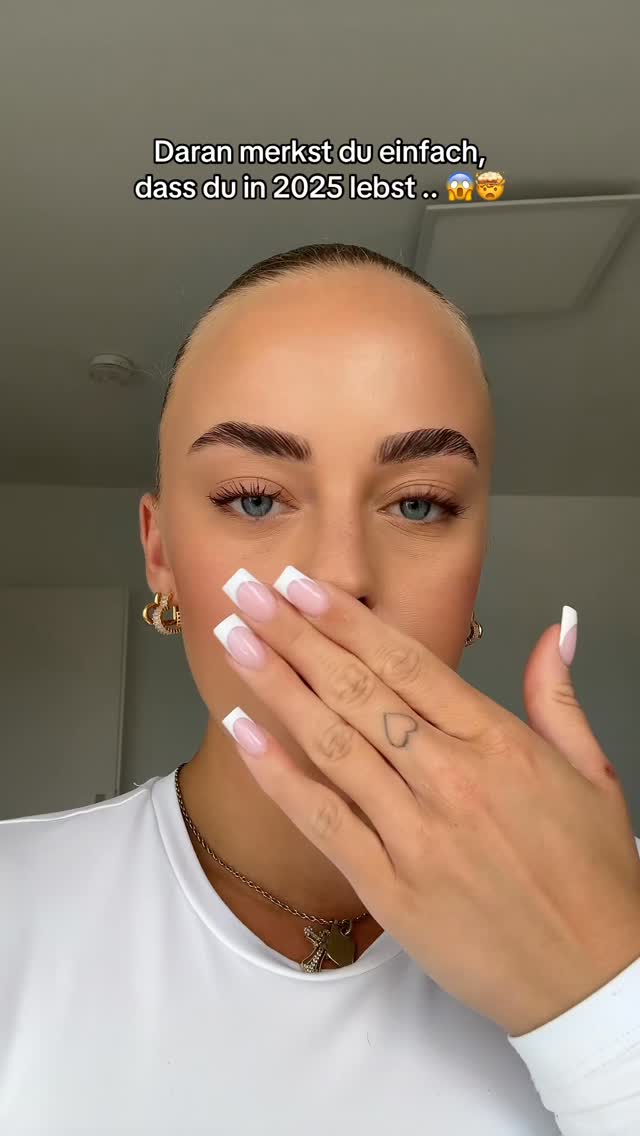 A close-up of a woman's face with light skin and light brown hair. Her eyes are bright blue, and she is partially covering her mouth with her right hand, revealing long, manicured nails with a French tip design. She wears gold hoop earrings and a white top. The background is a neutral color, and text overlaying the image reads, 'Daran merkst du einfach, dass du in 2025 lebst ...'.