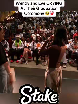 The image shows a candid moment at a graduation ceremony, with a blurred background of an auditorium filled with people. In the foreground, a person is seen from behind, wearing a light-colored outfit and holding a phone, seemingly capturing the event. The focus is immediately drawn to the text overlay at the top of the image, which reads "Wendy and Eve CRYING At Their Graduation Ceremony" with crying and heart emojis. In the bottom, the logo "Stake".