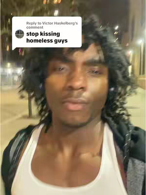 A selfie-style shot of a young Black man walking outdoors at night. He's wearing a white tank top and a black jacket. A speech bubble with white text, reading "stop kissing homeless guys," is superimposed on the image near the top left corner. The background is blurry, with visible streetlights.