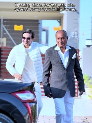 Two men, one in a white quilted jacket and sunglasses posing near a car, the other in a pinstriped jacket and white pants, are standing near a car in a well-lit outdoor setting. Text overlay at the top, 'Opening doors for the man who opened every door in life for me.'