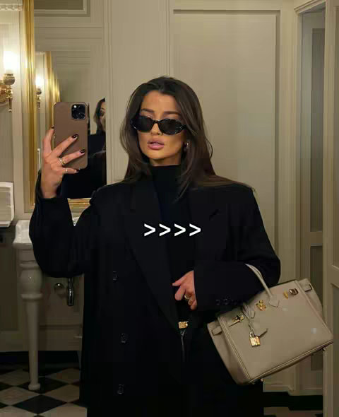 A woman is taking a selfie in a well-lit bathroom with a large mirror and a fancy cream-colored purse. She is wearing sunglasses and a black outfit, accessorized with jewelry. The background has a classic design.