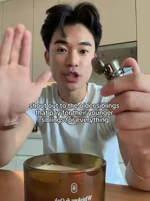A young man with dark hair is centered in the frame, holding a metallic object in his right hand and gesturing with his left. The background shows modern kitchen fixtures and a partially visible candle. Text overlays read, 'Shout out to the older siblings that pay for their younger siblings for everything'.