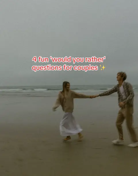 A couple is holding hands and smiling as they walk on a sandy beach. Text on the middle of the image says "4 fun 'would you rather' questions for couples". The background contains the ocean and an overcast sky.